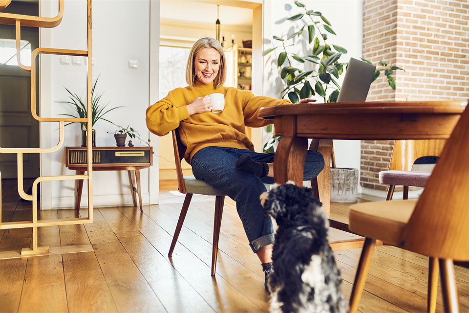 Entspannte Frau sitzt am Esstisch, lächelt ihren Hund an und arbeitet zu Hause am Computer.