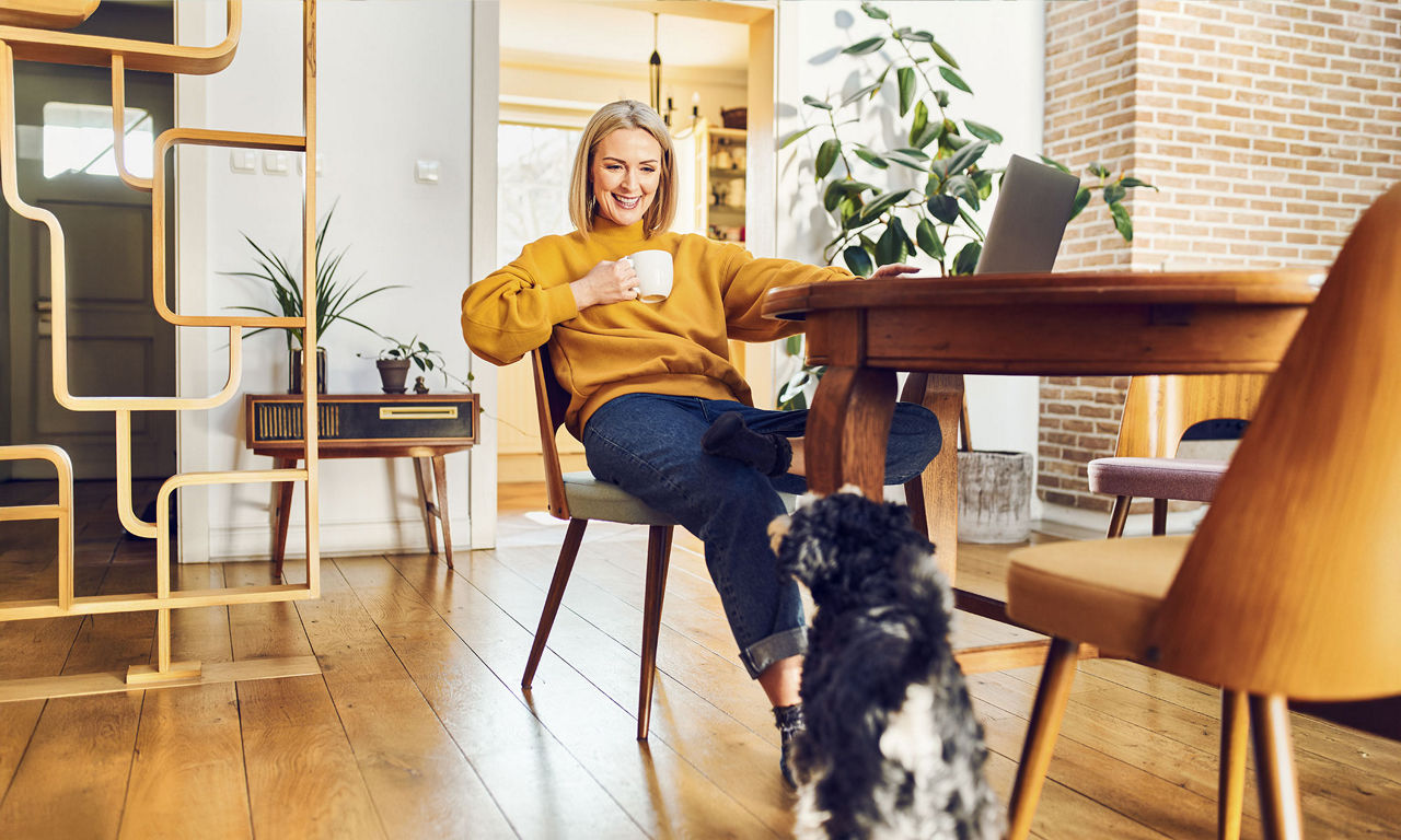 Entspannte Frau sitzt am Esstisch, lächelt ihren Hund an und arbeitet zu Hause am Computer.