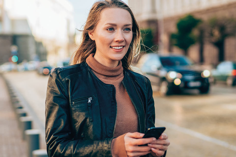 Junge Frau mit Smartphone in der Hand steht auf der Straße und lächelt.