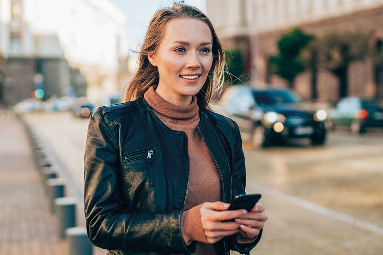 Junge Frau mit Smartphone in der Hand steht auf der Straße und lächelt