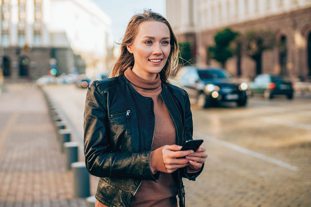 Junge Frau mit Smartphone in der Hand steht auf der Straße und lächelt