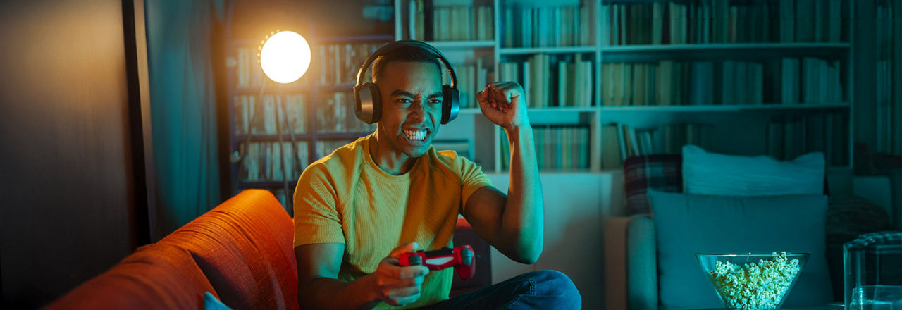 Junger Mann sitzt mit Kopfhörern auf dem Kopf  und Controller in der Hand auf einem Sofa 