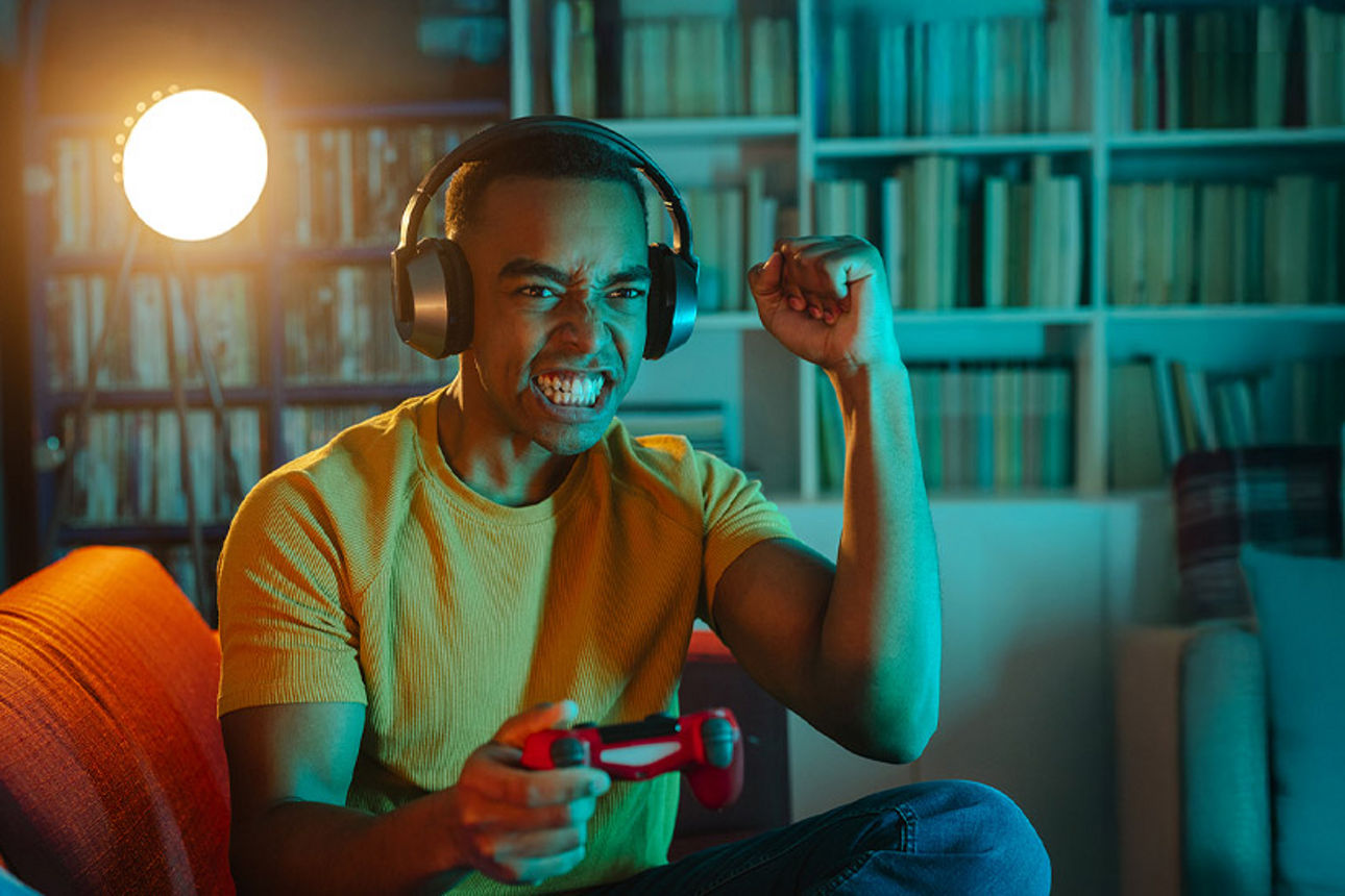 Junger Mann sitzt mit Kopfhörern auf dem Kopf  und Controller in der Hand auf einem Sofa 