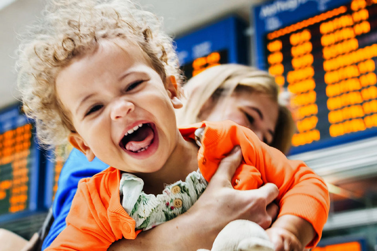 Junge Frau hält ihr lachendes Kind in den Armen, im Hintergrund eine Abflugtafel am Flughafen