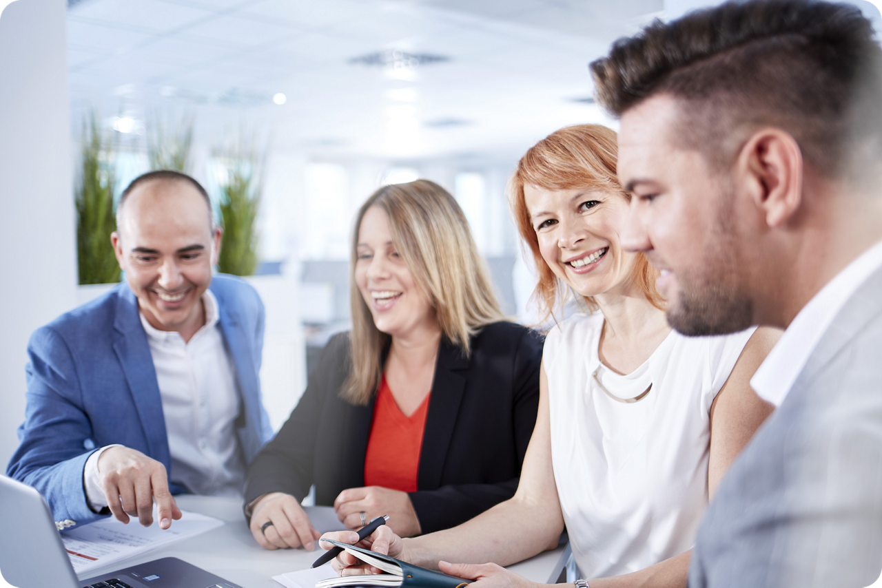 Zwei Männer und zwei Frauen stehen an einem Tisch und unterhalten sich, eine Frau lacht in die Kamera