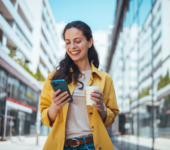 Eine junge Frau in einer gelben Jacke hat ein Smartphone und einen Kaffeebecher in der Hand und schaut glücklich aus