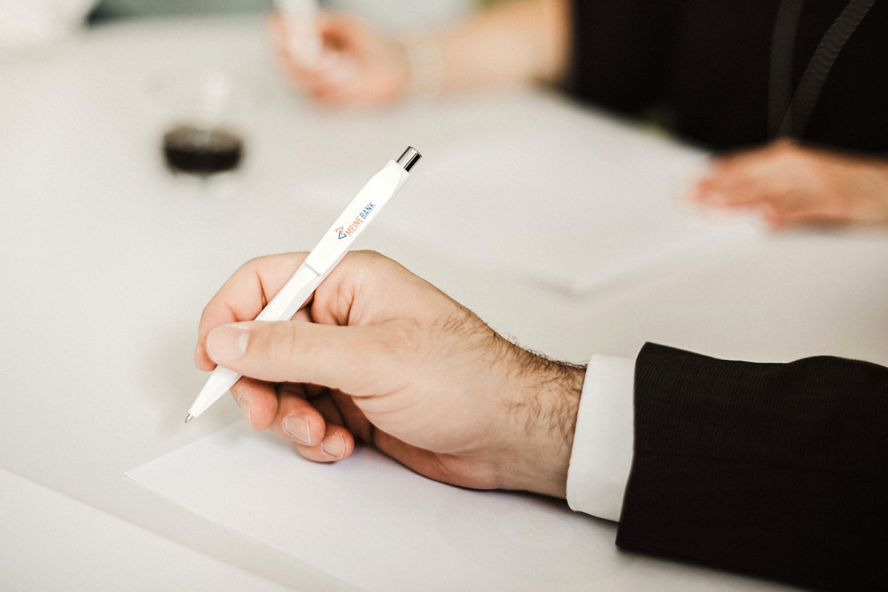 Abbildung zeigt eine Männerhand, die einen Kugelschreiber von MEINE BANK hält.