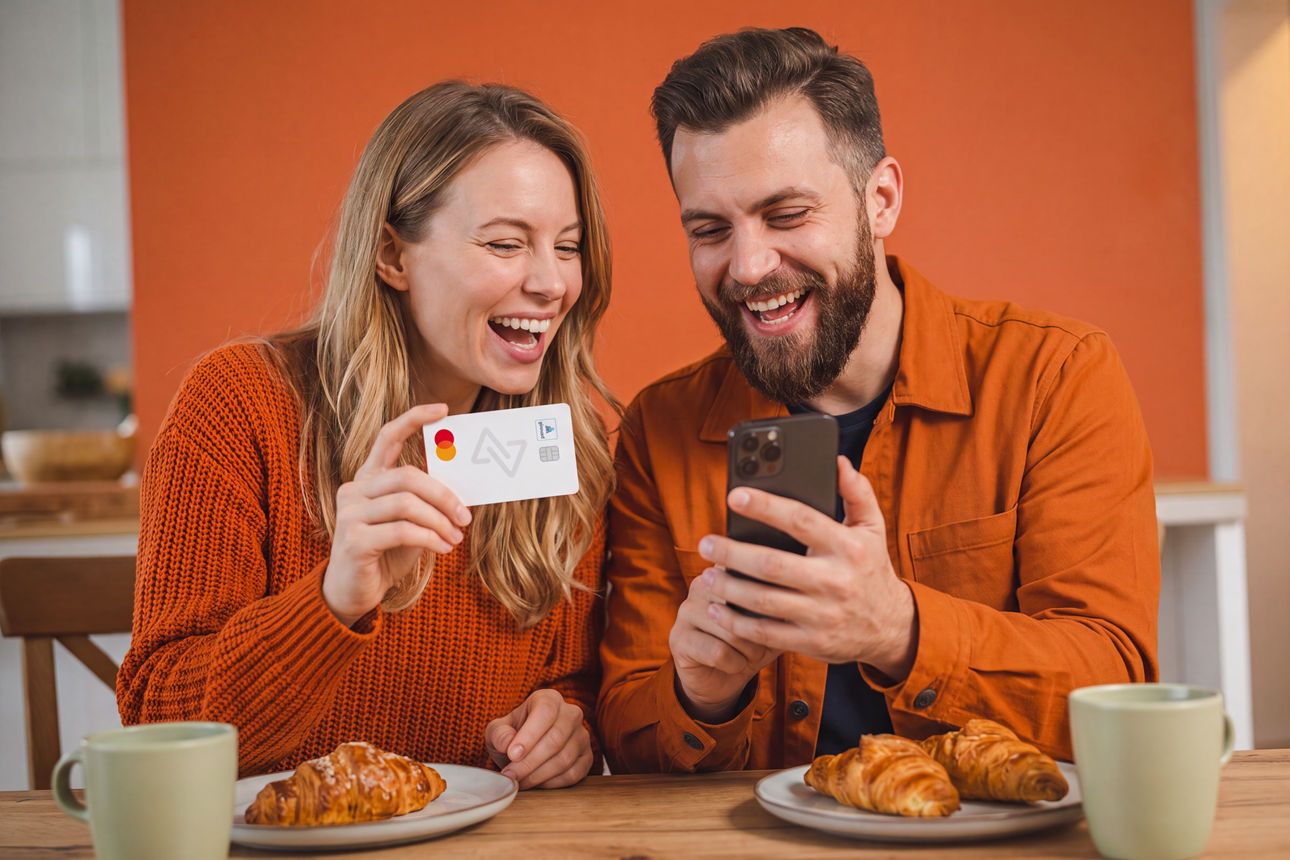 Abbildung zeigt ein fröhliches Paar am Frühstückstisch, das gemeinsam online einkauft und dabei die MEINE BANK girocard hält.
