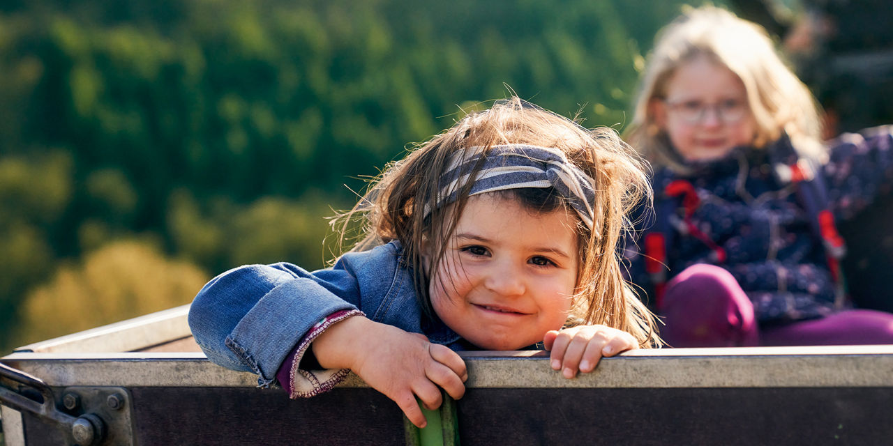 Zwei Kinder sitzen auf einer Anhängerplattform, wobei das vordere Kind lächelnd in die Kamera schaut