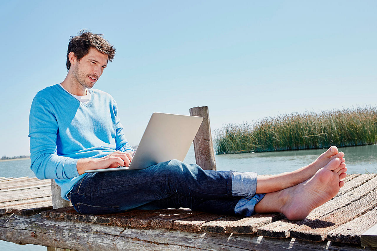 Ein Mann sitzt mit einem Laptop auf dem Schoß auf einem Steg