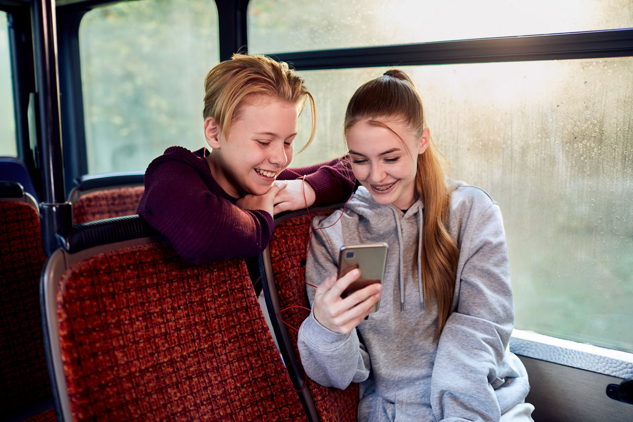 Zwei Jugendliche sitzen im Bus und schauen auf ein Handy.
