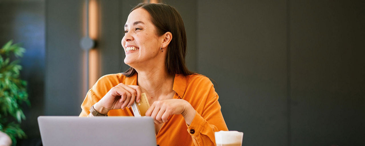 Eine Frau sitzt mit Laptop und Kaffee an einem Tisch und hält ihre goldene girocard in den Händen