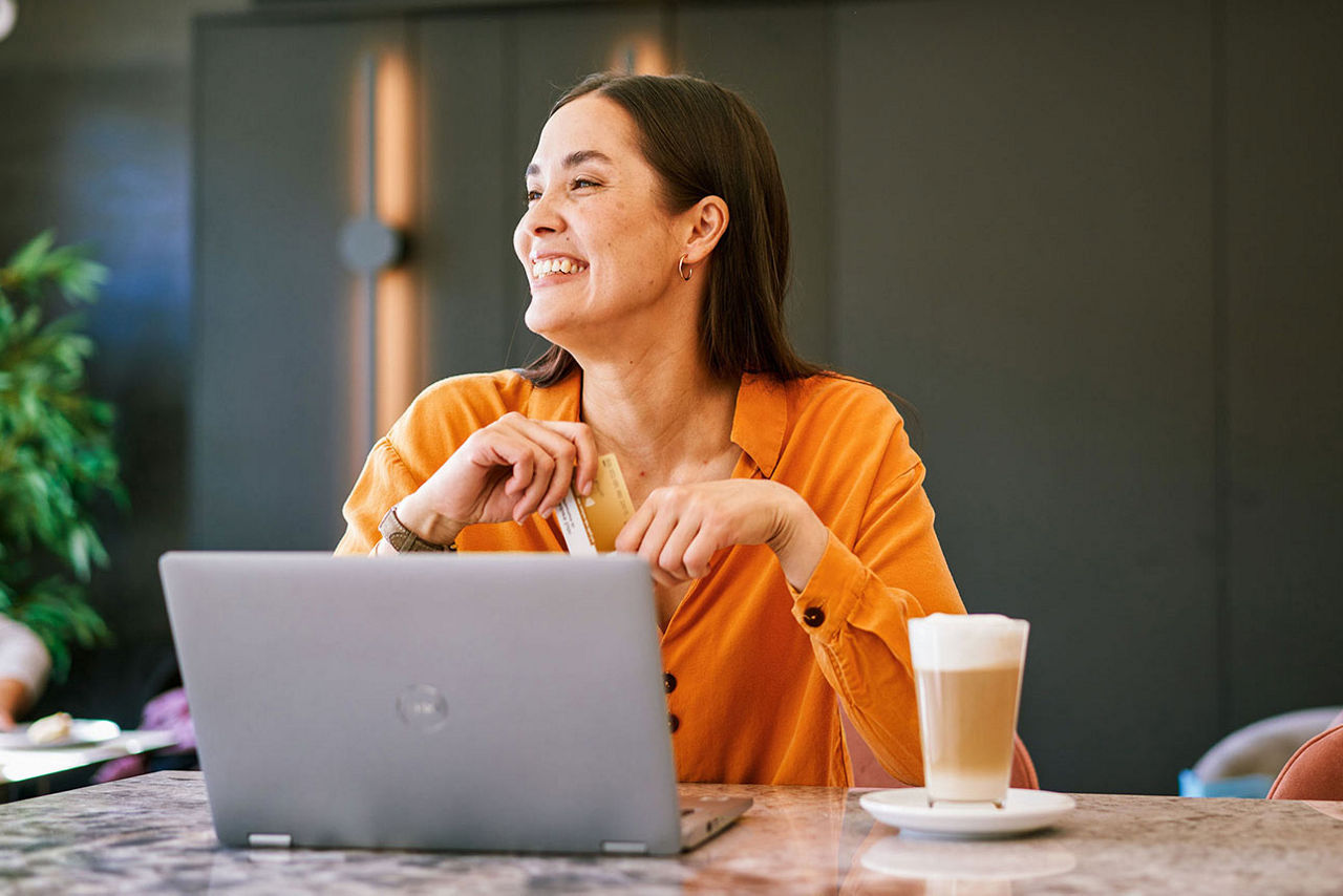 Eine Frau sitzt mit Laptop und Kaffee an einem Tisch und hält ihre goldene girocard in den Händen