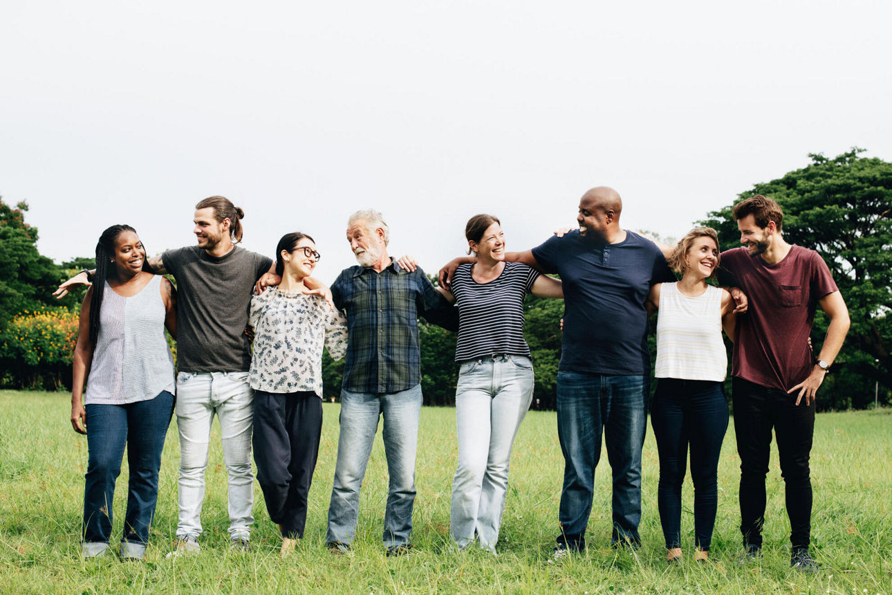 Unterschiedliche Menschen stehen im Park in einer Reihe, umarmen sich und lächeln sich an.