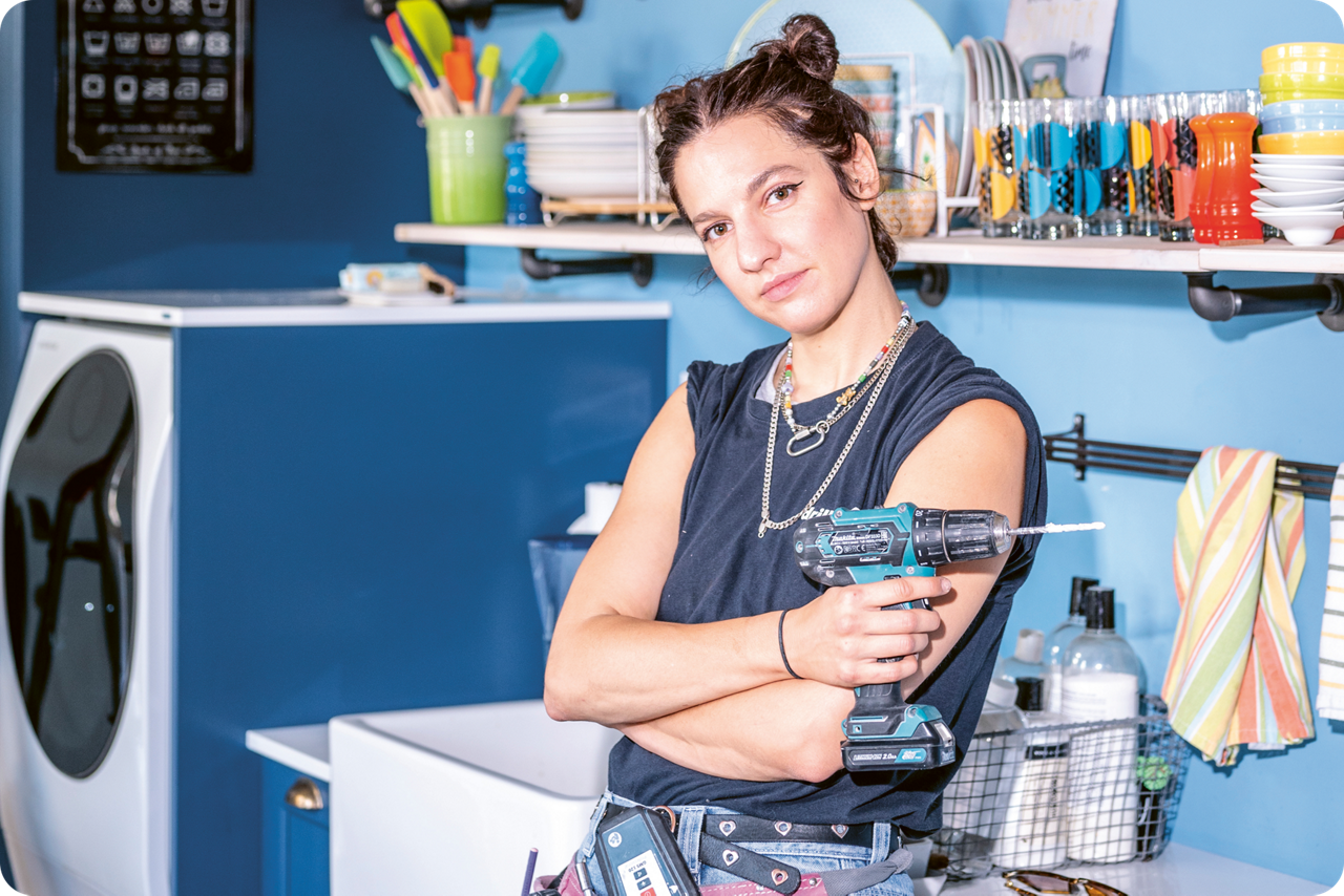 Eine Frau steht mit verschränkten Armen vor einer Spüle und hat einen Akkuschrauber in der Hand