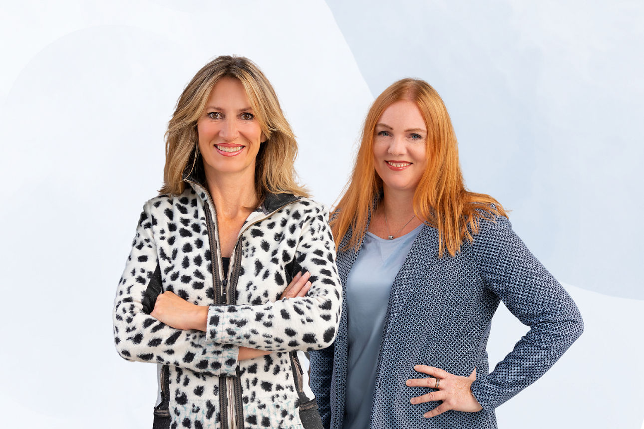 Zwei Frauen stehen nebeneinander, die Arme verschränkt, und schauen selbstbewusst in die Kamera.