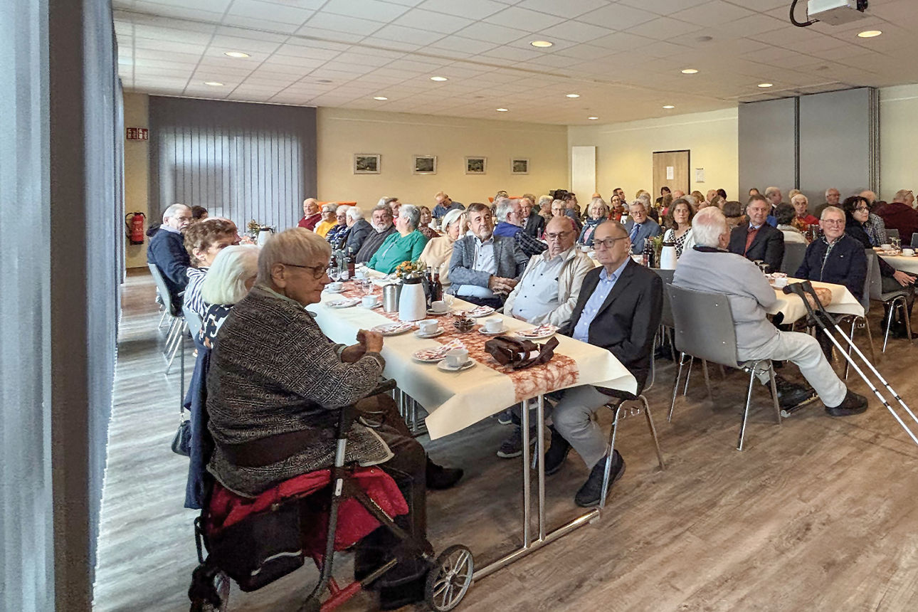 Personen sitzen an langen Tischreihen in einem größeren Raum mit Holzfußboden. Die Tische sind feierlich gedeckt.