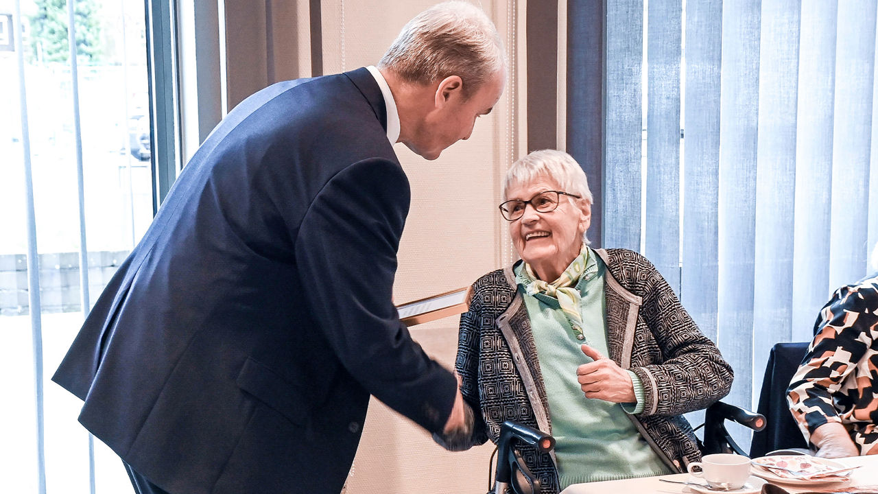 Konrad Breul schüttelt Marianne Kamps die Hand, die auf ihrem Rollator am Tisch sitzt und ihn herzlich anlächelt
