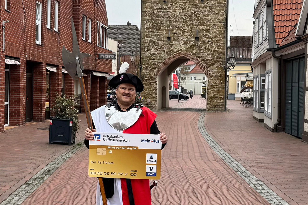 Ein Mann in mittelalterlicher Kleidung hält eine große, goldene Debitkarte vor sich.