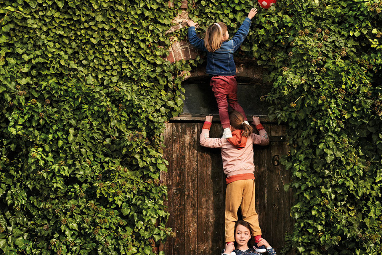 Drei Kinder bilden an einer eufeubewachsenen Wand eine Räuberleiter, um an einen Ball heran zu kommen