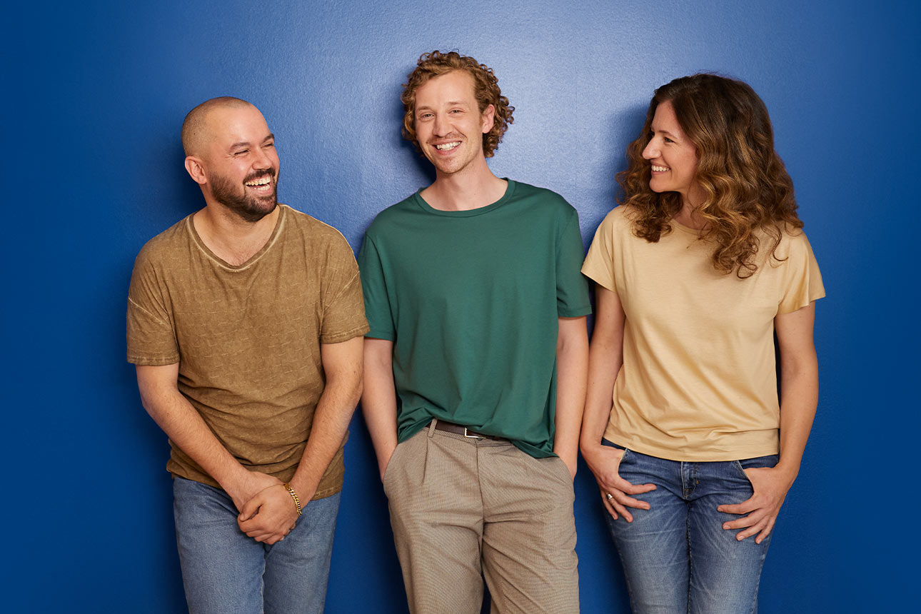 Drei lachende Menschen stehen vor einer blauen Wand mit dem Schriftzug „Wir sind meine Bank.”