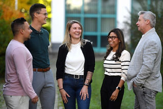 Eine Gruppe von fünf Erwachsenen stehen im Halbkreis zueinander. Davon sind zwei Frauen und drei Männer. Sie unterhalten sich und lachen dabei. Eine Gruppe von fünf Erwachsenen stehen im Halbkreis zueinander. Davon sind zwei Frauen und drei Männer. Sie unterhalten sich und lachen dabei.