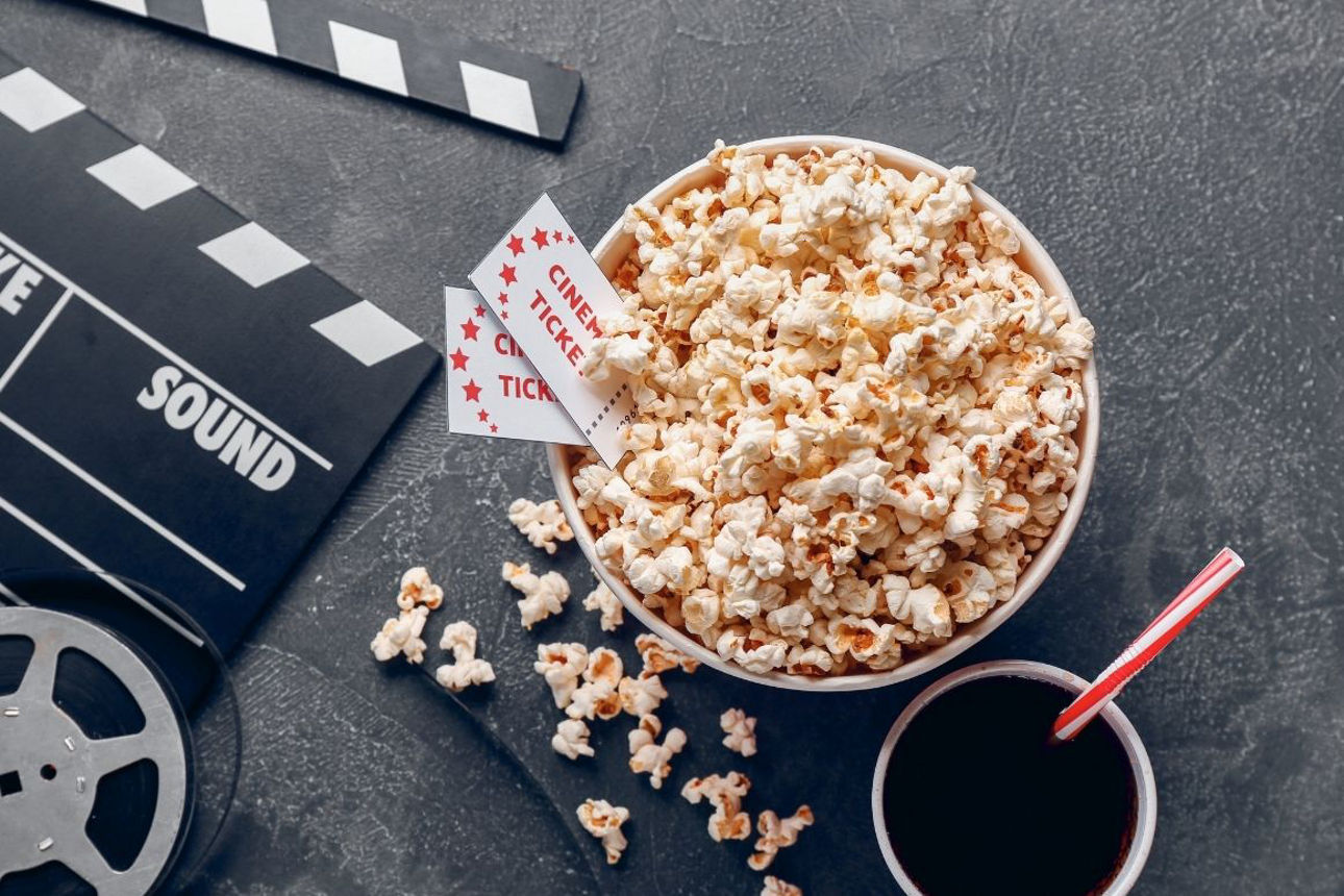 Kinoszene mit einer Schale Popcorn, zwei Kinokarten, einem Getränk sowie Filmklappe und Filmrolle auf dunklem Untergrund.