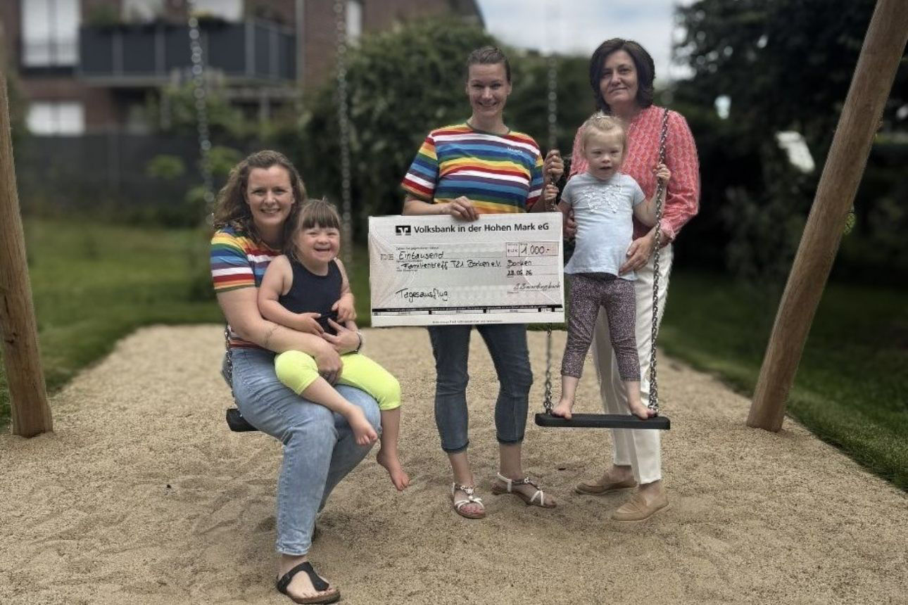 Vier Personen und zwei Kinder auf einem Spielplatz; zwei sitzen bzw. stehen an Schaukeln. Eine Person hält einen großen Scheck der Volksbank in der Hohen Mark eG über 1.000 Euro (Datum: 25.06.2026).