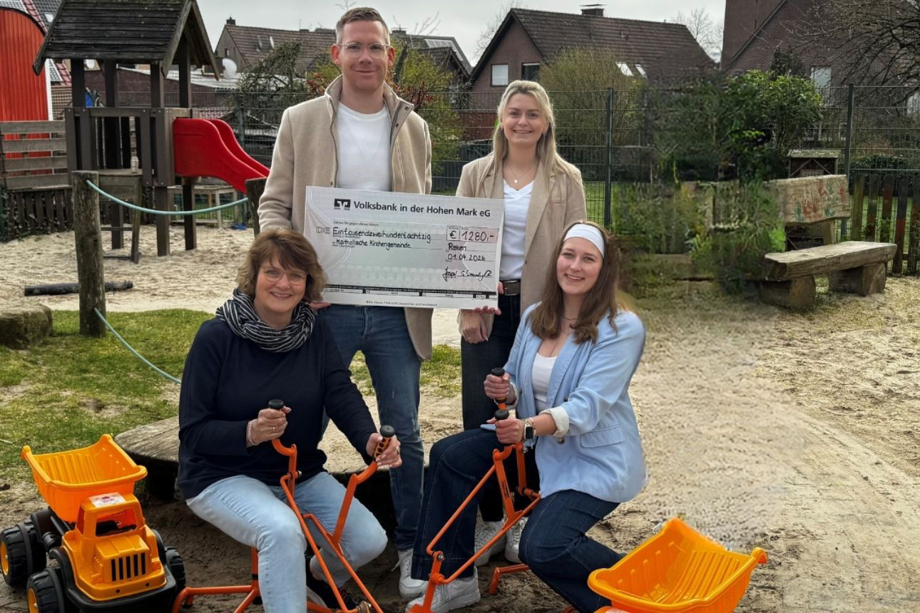 Vier Personen auf einem Spielplatz; zwei stehen, zwei sitzen auf Spielgeräten. In der Mitte wird ein Spendencheck der Volksbank in der Hohen Mark eG über 1.280 Euro an den Kitaverbund überreicht. Im Hintergrund sind Spielgeräte, Sandfläche und Wohnhäuser zu sehen.