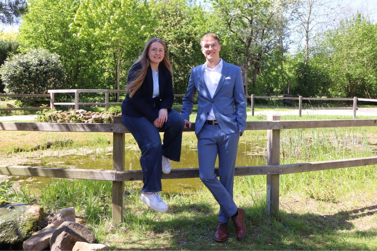 Zwei Personen lehnen an einem hölzernen Geländer vor einem kleinen Teich; im Hintergrund Bäume und Grünflächen in einer Park‑ oder Naturlandschaft.