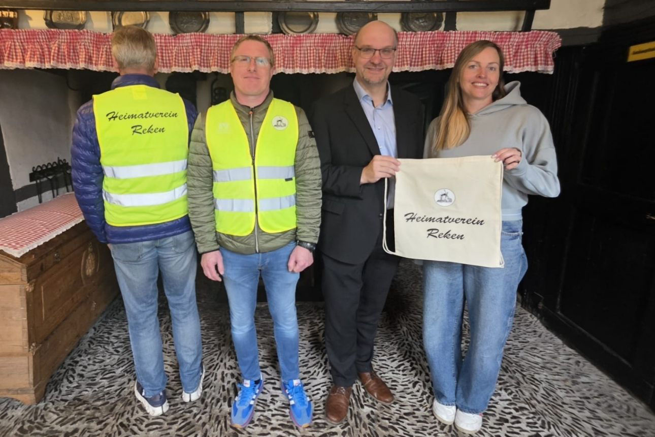 Vier Personen stehen in einem Innenraum nebeneinander. Zwei Personen tragen gelbe Warnwesten mit der Aufschrift „Heimatverein Reken“, eine davon zeigt die Rückseite. Eine Person in der Mitte hält gemeinsam mit einer weiteren Person einen hellen Stoffbeutel mit dem Aufdruck „Heimatverein Reken“. Im Hintergrund sind rustikale Holzmöbel und rot‑weiß karierte Stoffe zu sehen.