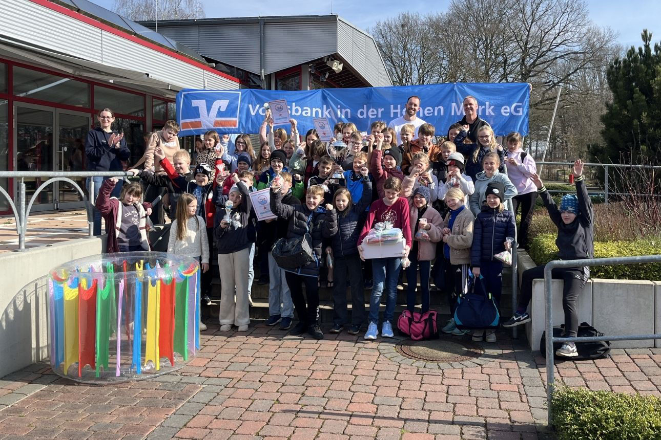 Auf dem Foto steht eine große Gruppe von Kindern und einigen Erwachsenen vor einem Gebäude im Außenbereich. Hinter der Gruppe hängt ein großes blaues Banner mit dem Logo und Namen der Volksbank in der Hohen Mark eG. Viele Kinder halten Urkunden oder kleine Preise in die Höhe, einige tragen Geschenkboxen. Die Gruppe wirkt fröhlich und feiernd. Im Vordergrund steht ein rundes, buntes Wasserspielgerät. Rechts und links sind Geländer, Pflanzen und ein gepflasterter Bereich zu sehen. Die Szene findet bei sonnigem Wetter statt.