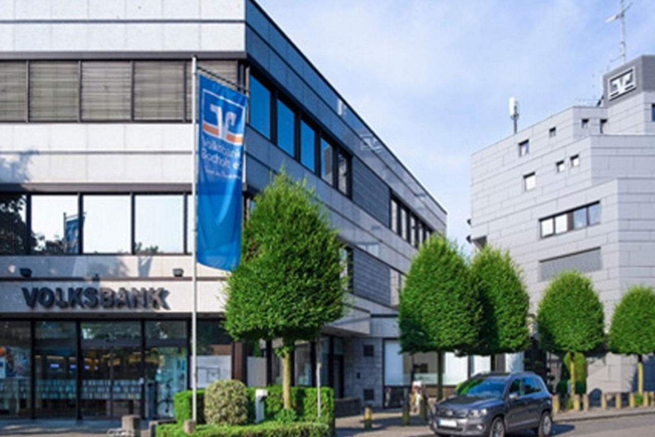 Zu sehen ist die Volksbank Bocholt: Ein modernes, mehrgeschossiges Bankgebäude mit hellgrauer Fassadenverkleidung und großen Fensterflächen. Am Haupteingang ist ein Volksbank‑Schriftzug angebracht, daneben hängt eine blaue Fahne mit Volksbank‑Logo. Entlang der Straße stehen regelmäßig geschnittene Bäume, davor ist ein geparktes Auto zu sehen. Im Hintergrund befindet sich ein weiteres Gebäude mit Volksbank‑Logos.