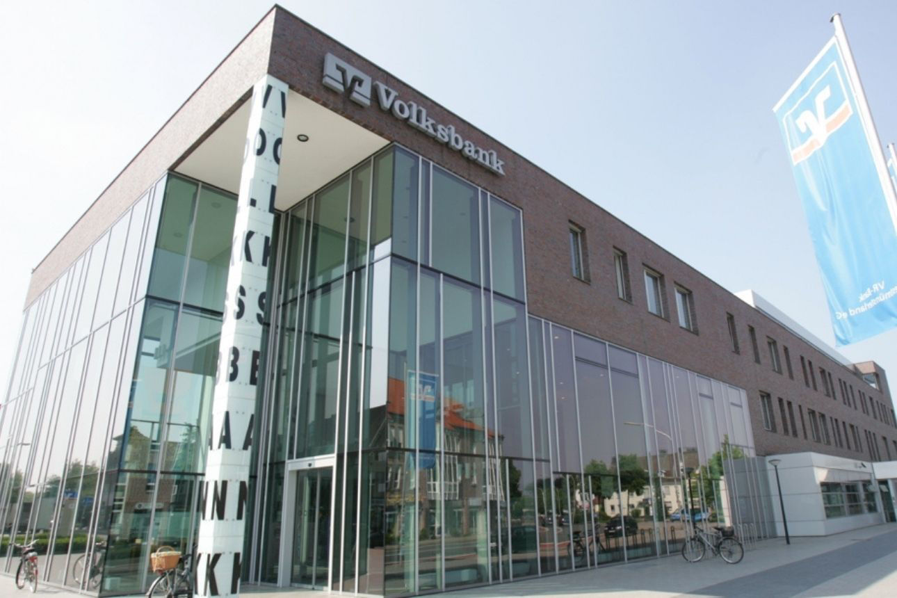 Zu sehen ist die Volksbank Westmünsterland eG mit einem modernen, mehrgeschossiges Volksbank‑Gebäude mit einer Kombination aus Backstein- und Glasfassade. Der Eingangsbereich ist vollständig verglast, darüber befindet sich ein Volksbank‑Schriftzug. Vor dem Gebäude stehen Fahrräder, rechts ist eine blaue Fahne mit Volksbank‑Logo angebracht.