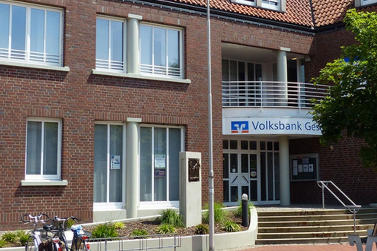 Zweigeschossiges Backsteingebäude mit rotem Ziegeldach. Vor dem Eingang führen Stufen zu einer Glastür mit überdachtem Balkon, darüber ist ein Volksbank‑Gescher‑Schriftzug angebracht. Links stehen mehrere Fahrräder, rechts grenzt ein begrünter Bereich an das Gebäude.