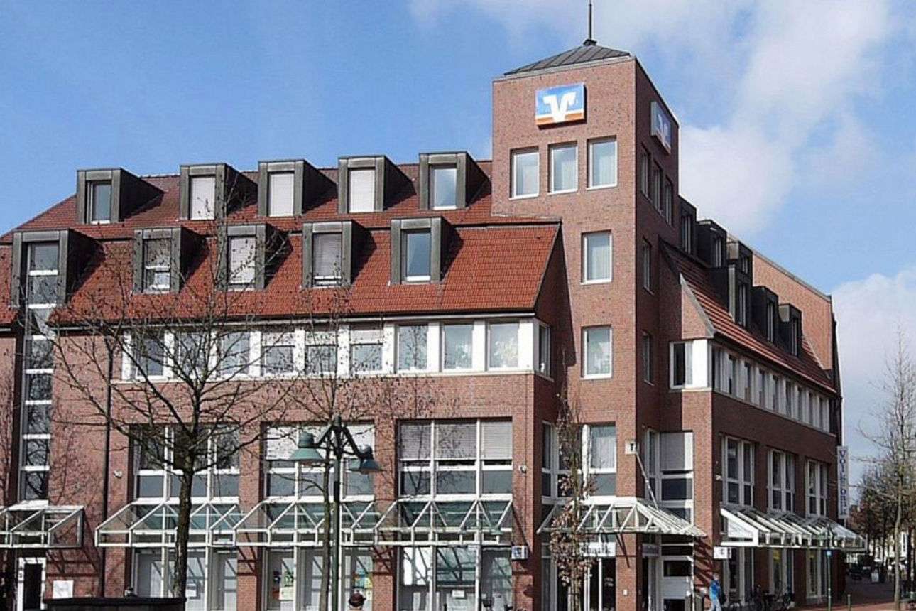 Zu sehen ist die Volksbank Gronau-Ahaus. Ein Mehrgeschossiges Backsteingebäude mit rotem Ziegeldach und einem markanten Eckturm. An der Fassade des Turms ist ein Volksbank‑Logo angebracht. Im Erdgeschoss befinden sich große Fenster und gläserne Vordächer über den Eingängen. Vor dem Gebäude verläuft eine Fußgängerzone mit einzelnen Passanten und Bäumen.