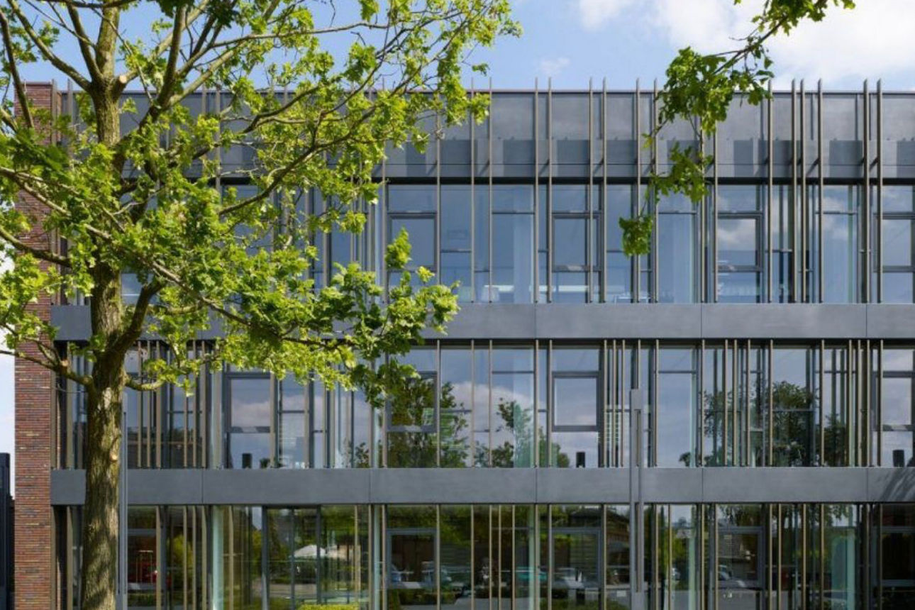 Zu sehen ist die Volksbank Raesfeld-Erle: Eine Luftaufnahme eines mehrteiligen Backsteingebäudes mit roten Ziegeldächern. Mehrere Dachflächen mit Gauben und Dachfenstern sind sichtbar. Das Gebäude steht an einer Straßenecke, angrenzend befinden sich weitere Gebäude und Bäume.