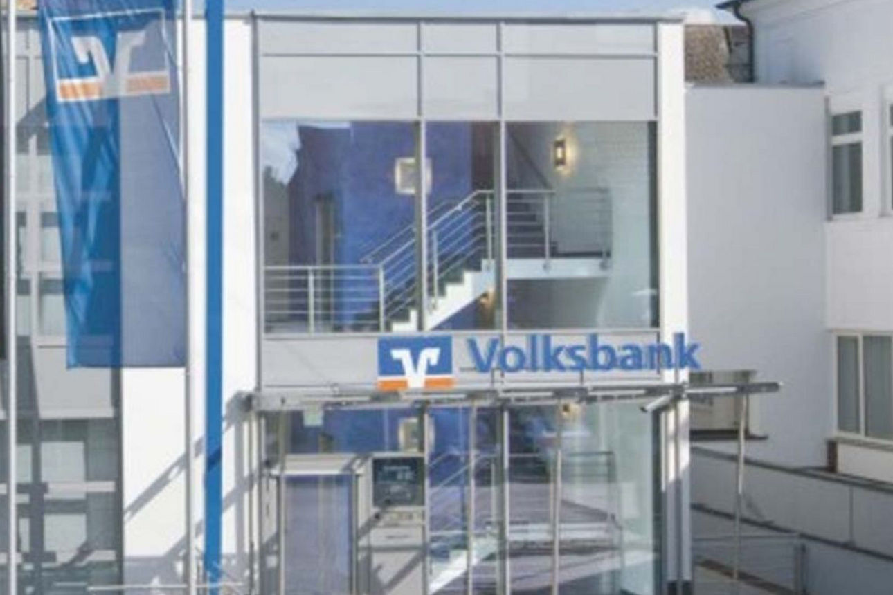 Das Bild zeigt die Volksbank Ochtrup-Laer mit einem modernen Bankgebäude mit weißer Fassade und großem Glas-Eingangsbereich. Über dem Eingang ist ein Volksbank‑Schriftzug mit Volksbank‑Logo angebracht. Durch die Glasfront sind eine Treppe und das Innere des Gebäudes sichtbar. Links vor dem Gebäude hängen Fahnen, darunter eine Deutschlandflagge.
