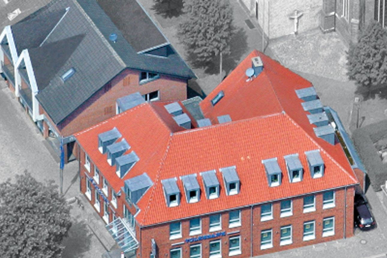 Zu sehen ist die Volksbank Raesfeld-Erle: Eine Luftaufnahme eines mehrteiligen Backsteingebäudes mit roten Ziegeldächern. Mehrere Dachflächen mit Gauben und Dachfenstern sind sichtbar. Das Gebäude steht an einer Straßenecke, angrenzend befinden sich weitere Gebäude und Bäume.