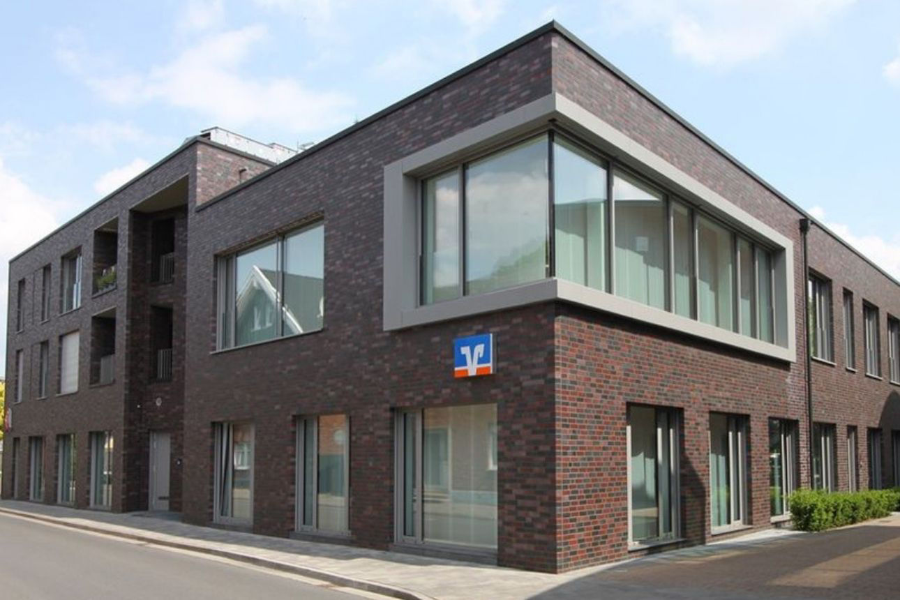 Zu sehen ist die Volksbank Rhede: Ein modernes, zweigeschossiges Gebäude aus dunklem Backstein an einer Straßenecke. Große Fensterflächen um die Gebäudeecke prägen die Fassade, darunter befindet sich ein Volksbank‑Logo. Entlang der Straße verlaufen Gehweg und Fahrbahn.