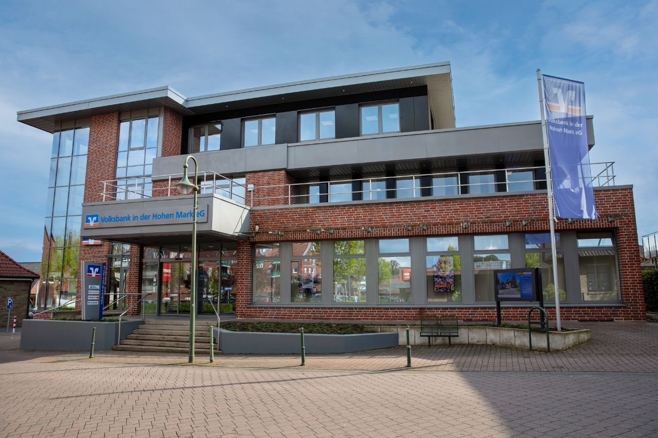 Zu sehen ist das Gebäude der Volksbank in der Hohen Mark eG: Ein mehrgeschossiges Gebäude aus Backstein mit grauen Fassadenelementen und großen Fensterflächen. Der Haupteingang ist leicht erhöht und mit einem Schild „Volksbank in der Hohen Mark eG“ gekennzeichnet. Rechts vor dem Gebäude steht eine Fahne mit Volksbank‑Logo, davor befindet sich ein gepflasterter Vorplatz.