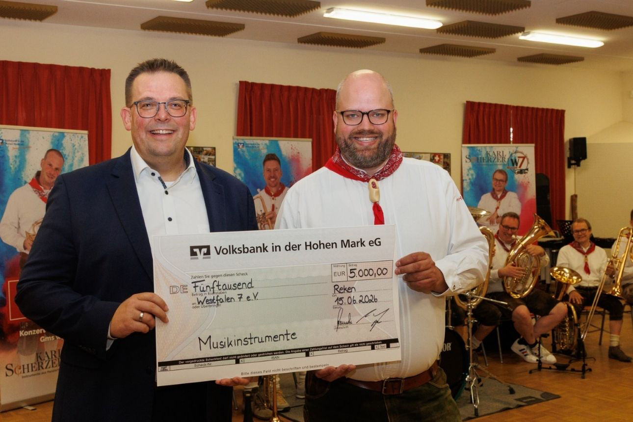 Zwei Personen halten gemeinsam einen großen Spendenscheck der Volksbank in der Hohen Mark eG über 5.000 Euro, ausgestellt an „Westfalen 7 e. V.“ für „Musikinstrumente“ (Datum: 15.06.2024, Ort: Reken). Im Hintergrund sitzt eine Blasmusikgruppe mit Instrumenten.