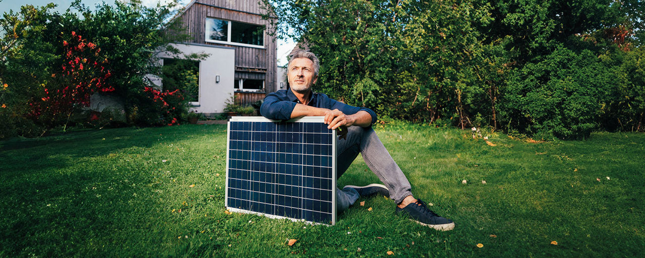 Ein Mann sitzt vor einem Haus im Garten und hält ein Solarpanel in den Händen.