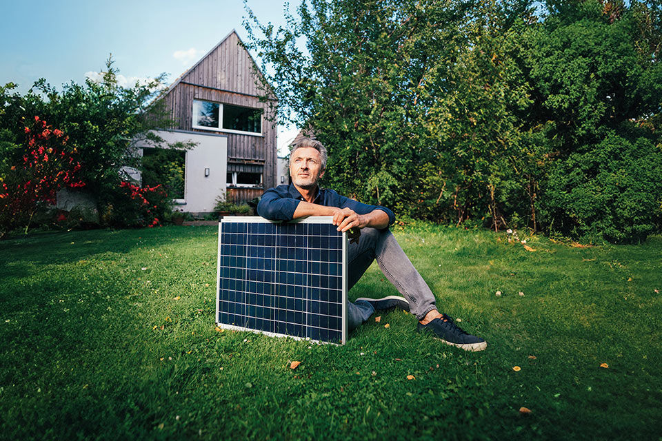 Ein Mann sitzt vor einem Haus im Garten und hält ein Solarpanel in den Händen.