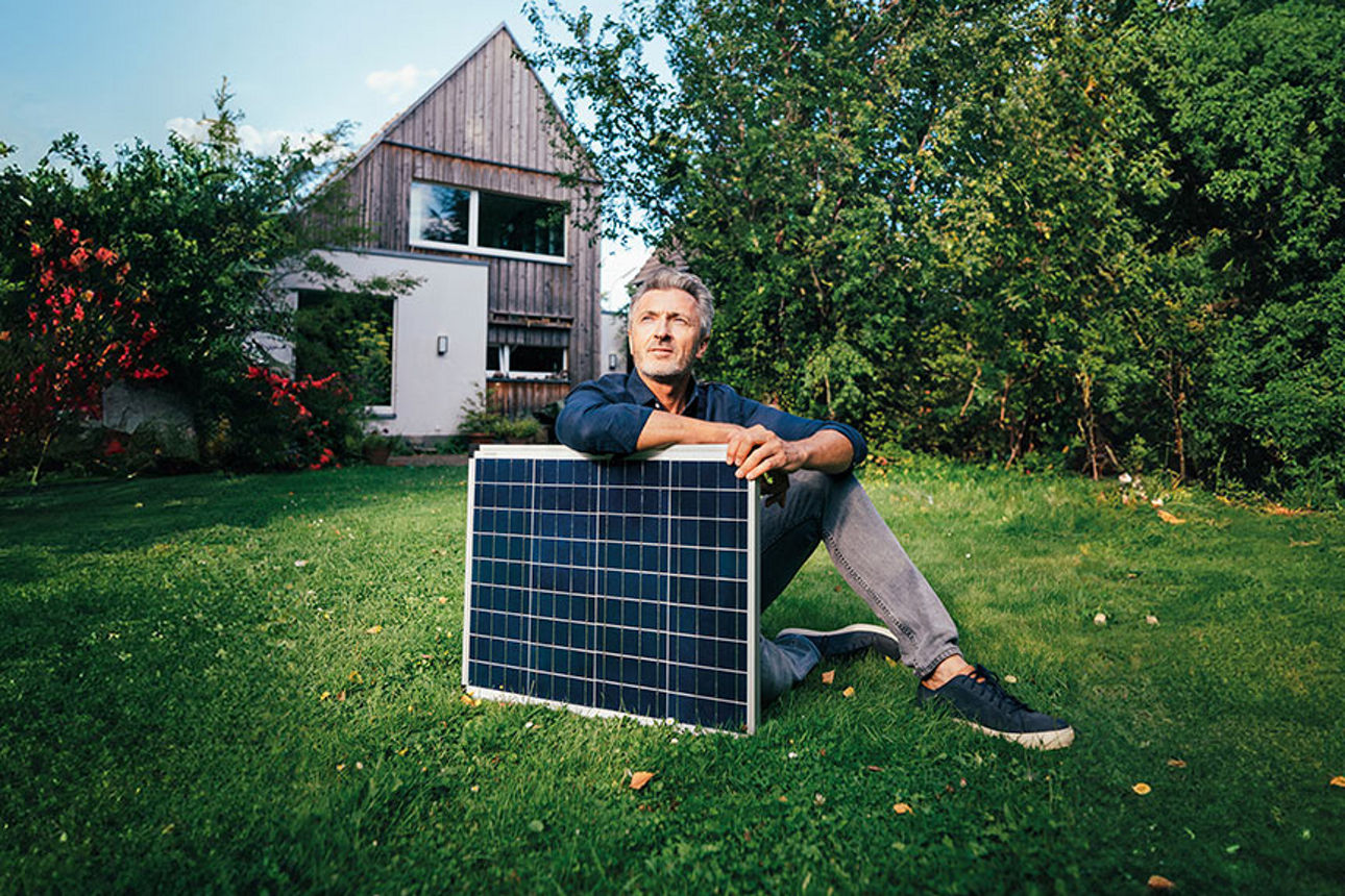 Mann sitzt mit einem Solarpanel in seinem Garten auf dem Rasen im Hintergrund sieht man sein Haus