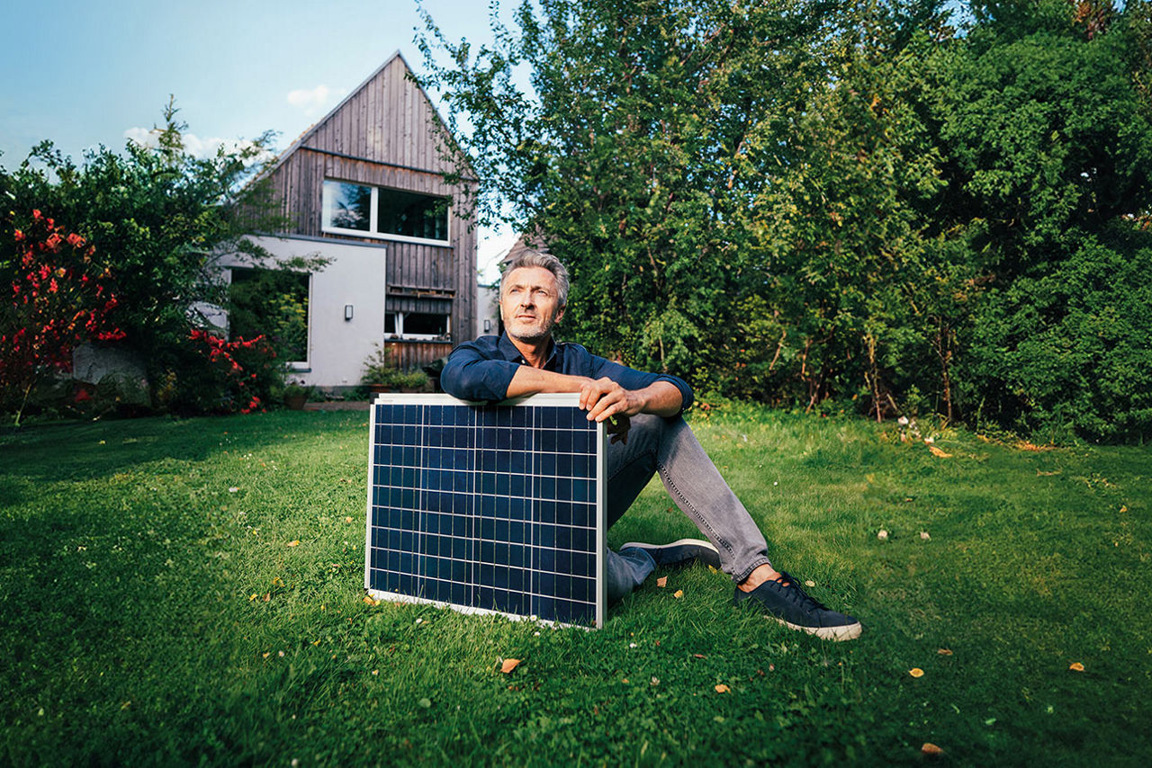 Ein Mann sitzt vor einem Haus im Garten und hält ein Solarpanel in den Händen.