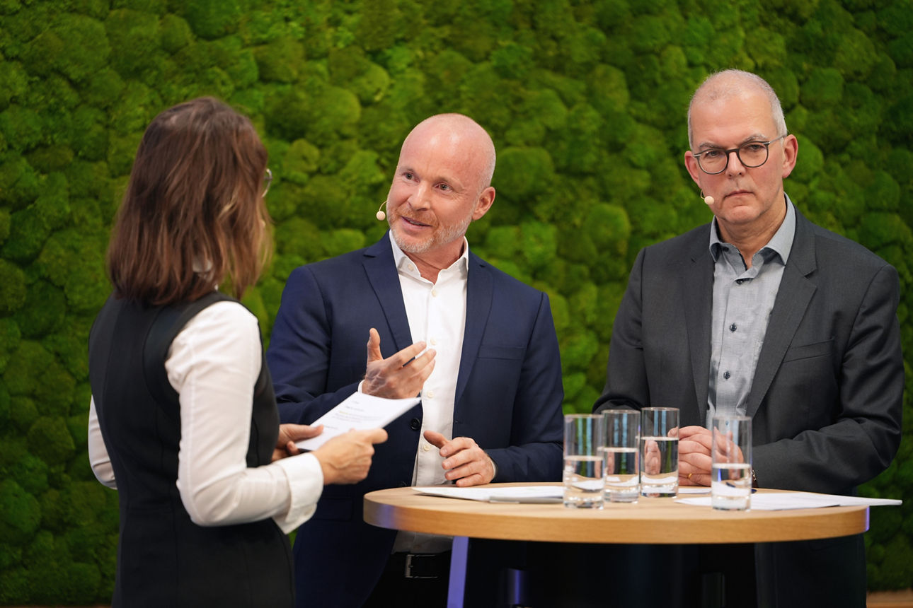 Ein Frau und zwei Männer im Austausch vor einer Mooswand