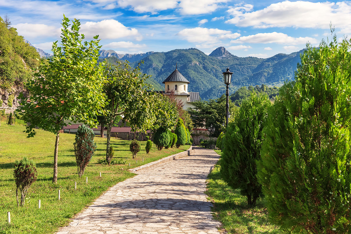 Moraca-Kloster in Montenegro: Ein hell gepflasterter Weg führt an Zypressen, Bäumen und Wiesen vorbei auf die Klostermauer zu