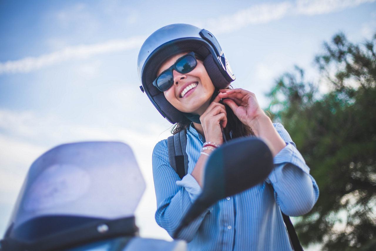 Eine Junge Frau steht neben ihrem Moped, wobon nur ein Teil des Lenkers zu sehen ist. Die Dame trägt eine Sonnebrille und einen Helm, den sie gerade schließt. 