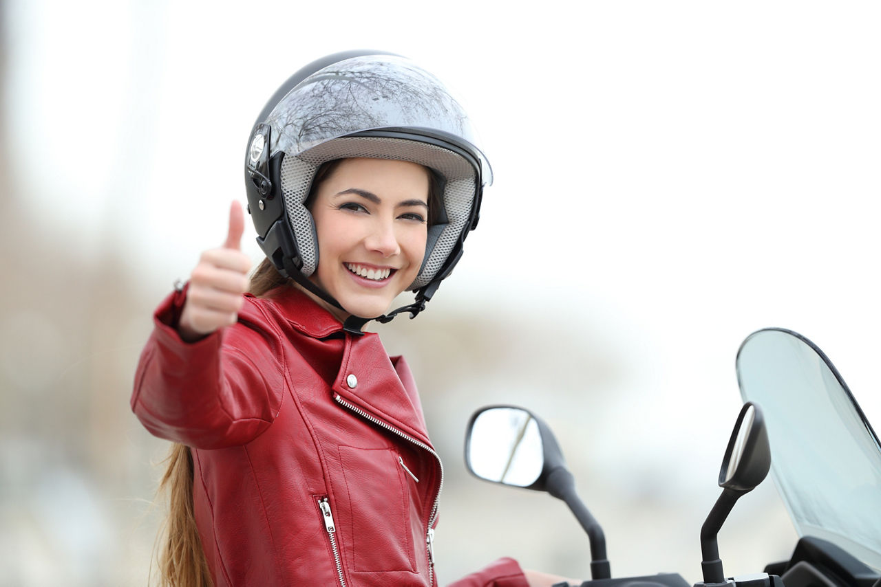 junge Frau sitzt mit einem Helm auf dem Kopf auf einem Roller oder Moped und hält ihren Daumen nach oben in die Kamera